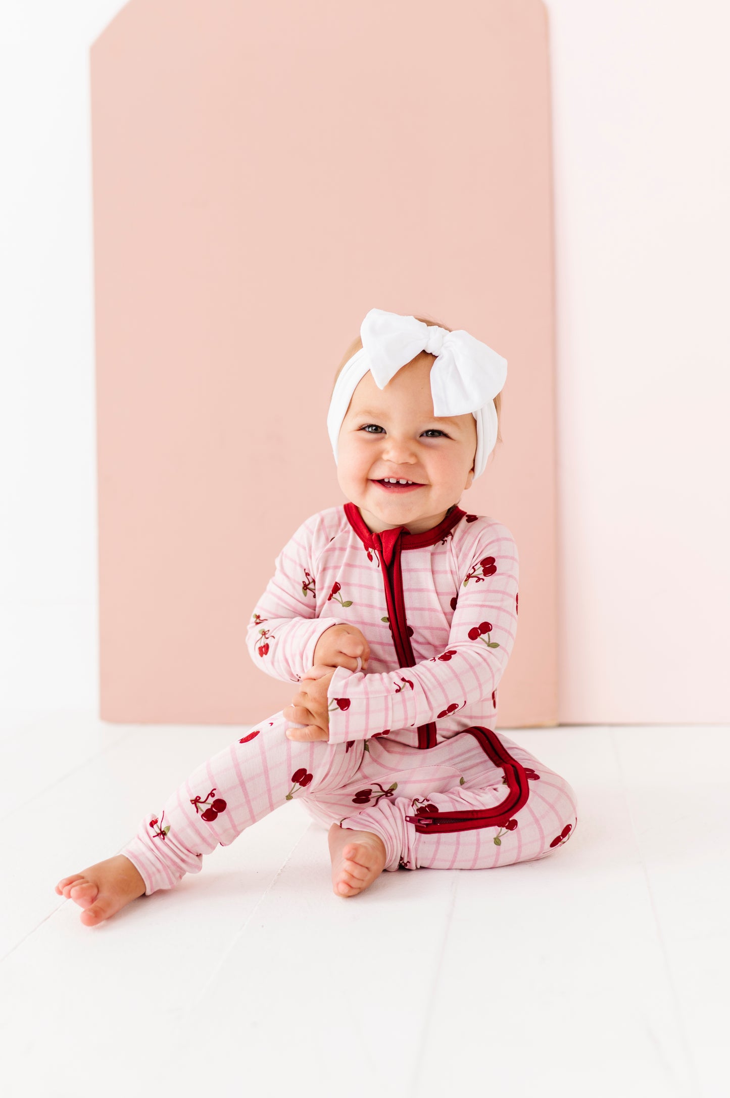 Baby in a pink outfit with red details and a white bow sitting on a white surface.