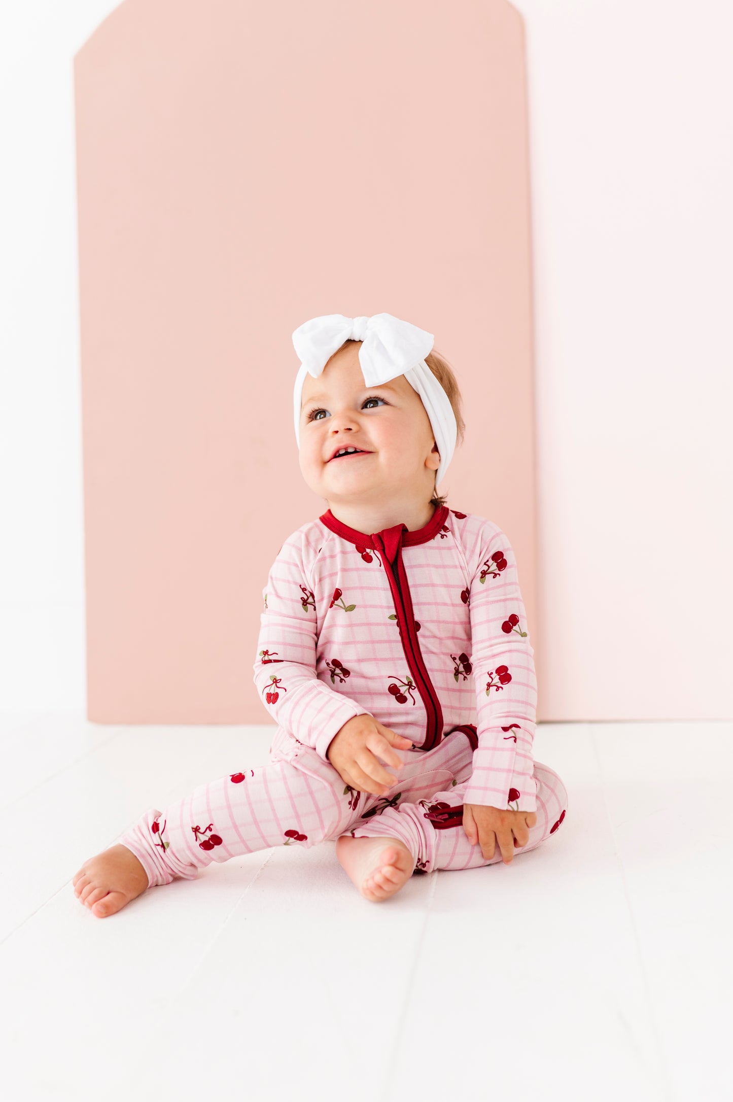 Baby wearing a pink outfit with red accents and a white headband, sitting against a light pink background.