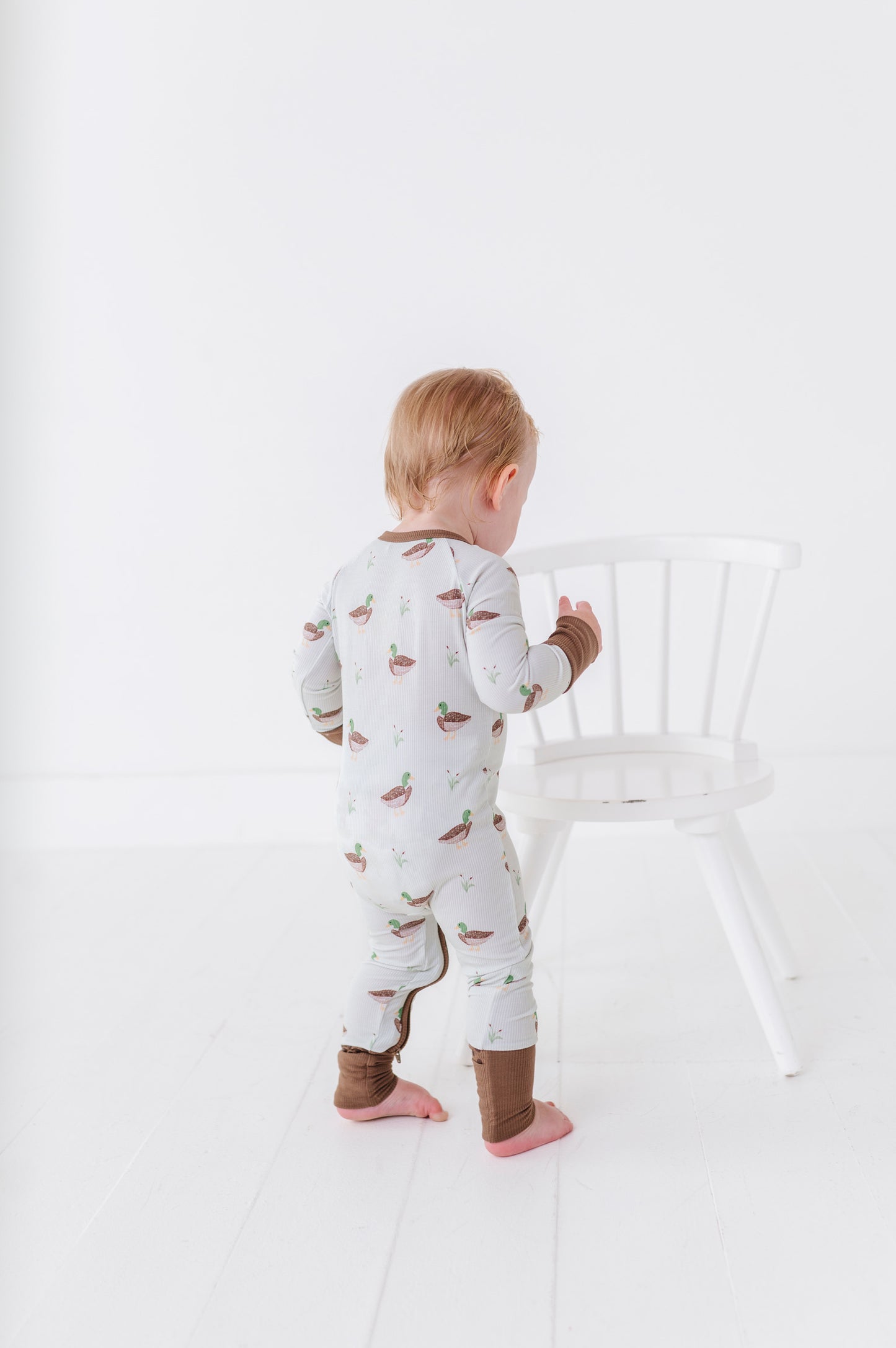Child wearing a patterned onesie standing next to a white chair on a plain background