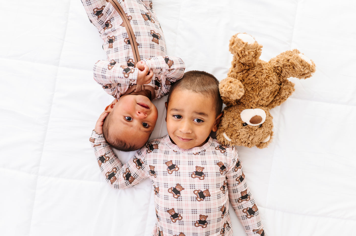 Two children in matching pajamas with teddy bear patterns on a white bed, one holding a teddy bear.