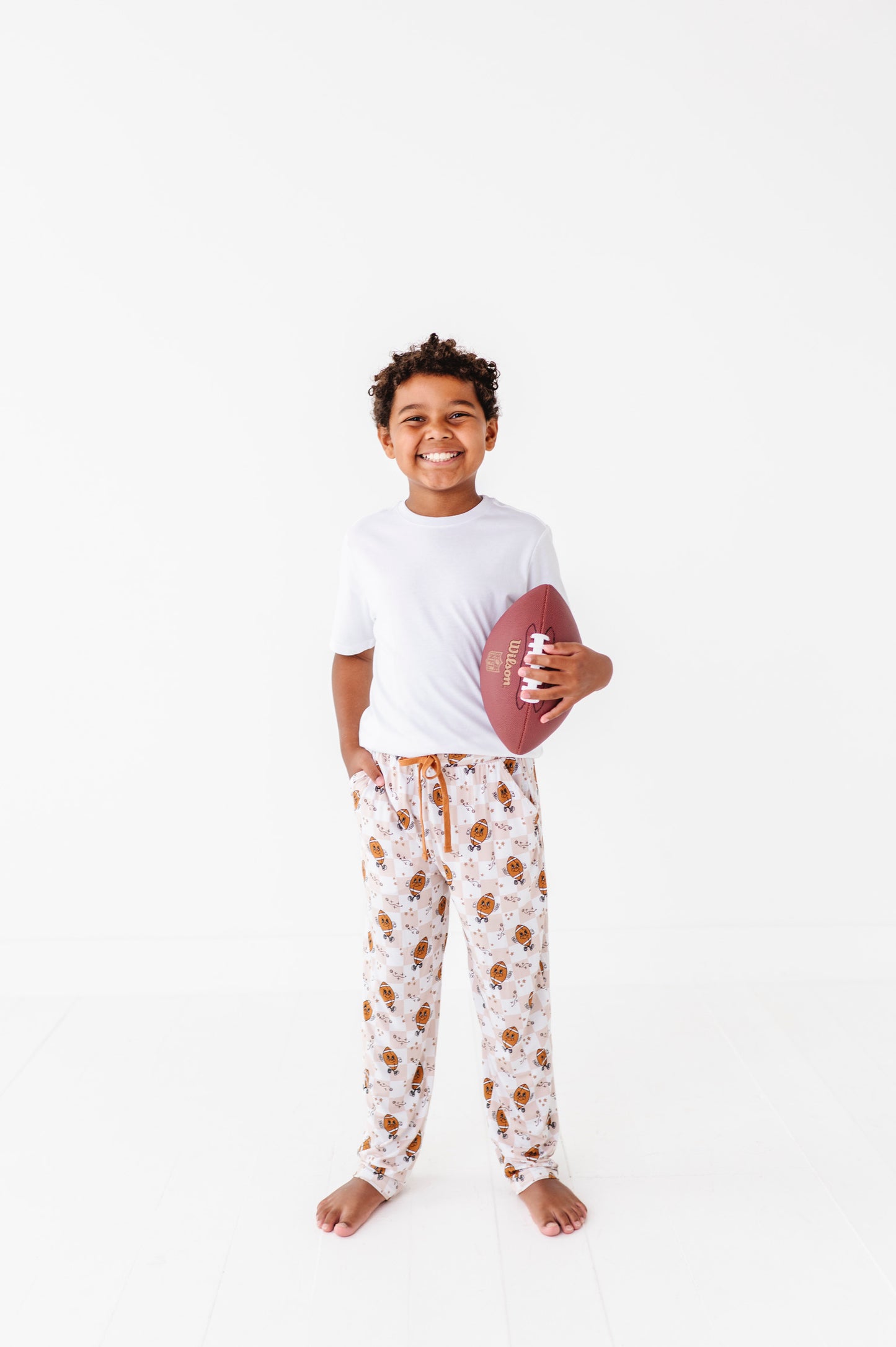 Child holding a football wearing a white t-shirt and patterned pajama pants on a white background