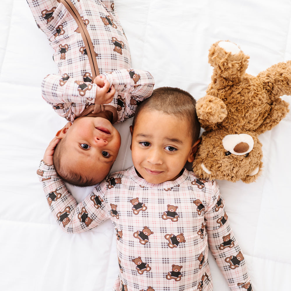 
                      
                        Two children in matching pajamas with teddy bear patterns on a white bed, one holding a teddy bear.
                      
                    